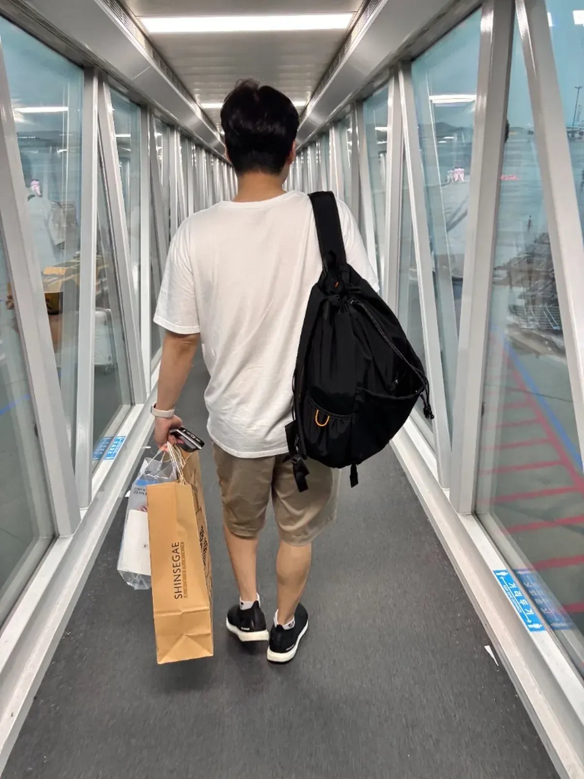 Traveler carrying Shinsegae duty-free shopping bags through an airport boarding bridge
