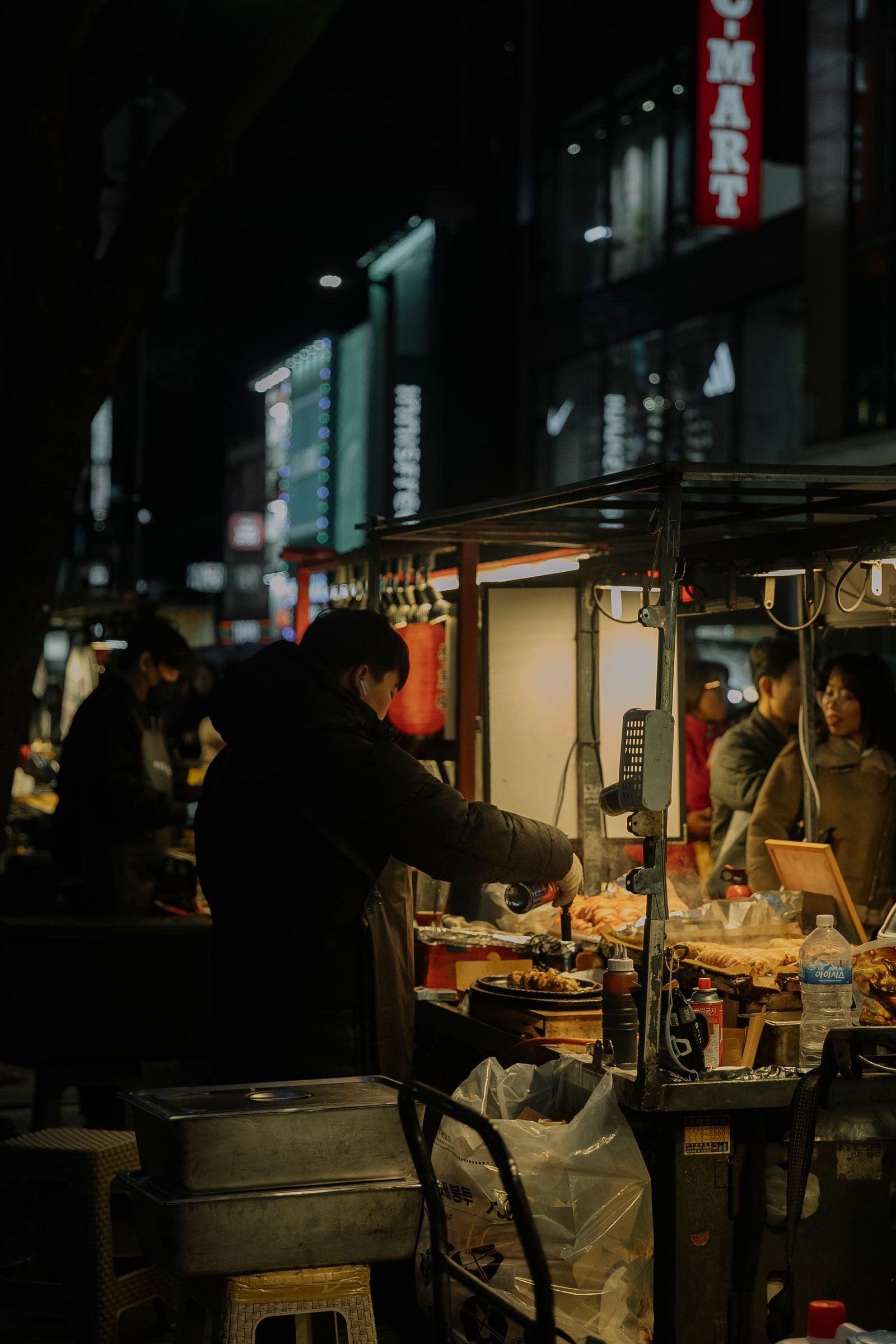 A vendor serves food at an illuminated street market in Seoul at night.