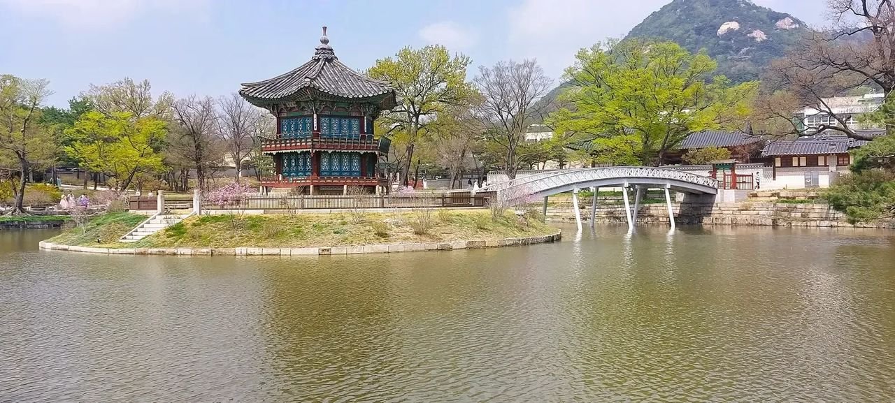 Hyangwonjeong Pavilion stands on a small island with a white arched bridge and trees around the pond.