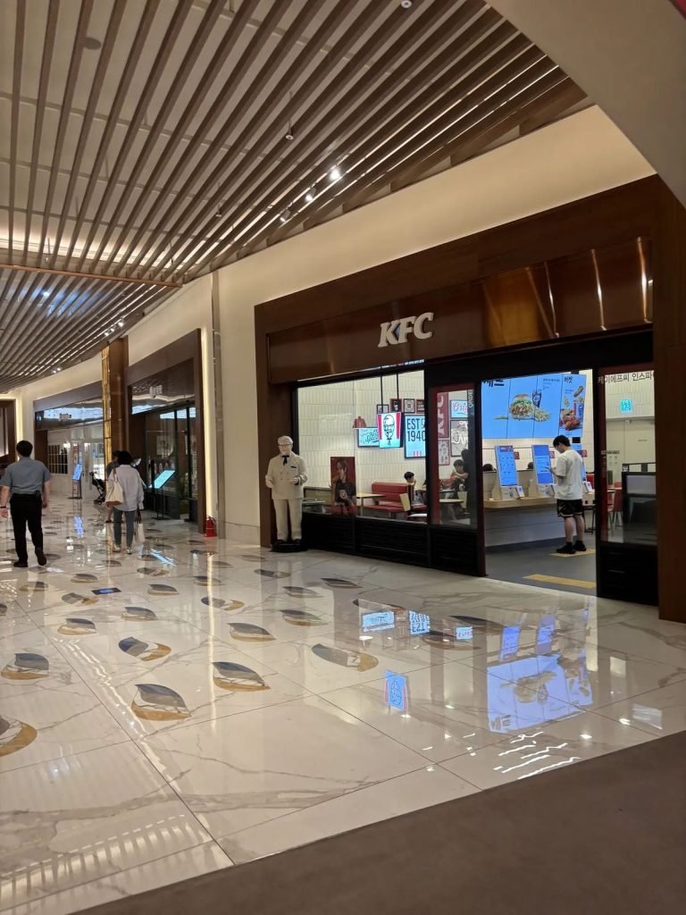Self-order kiosks inside a KFC restaurant in Korea, with a customer using one of the screens.