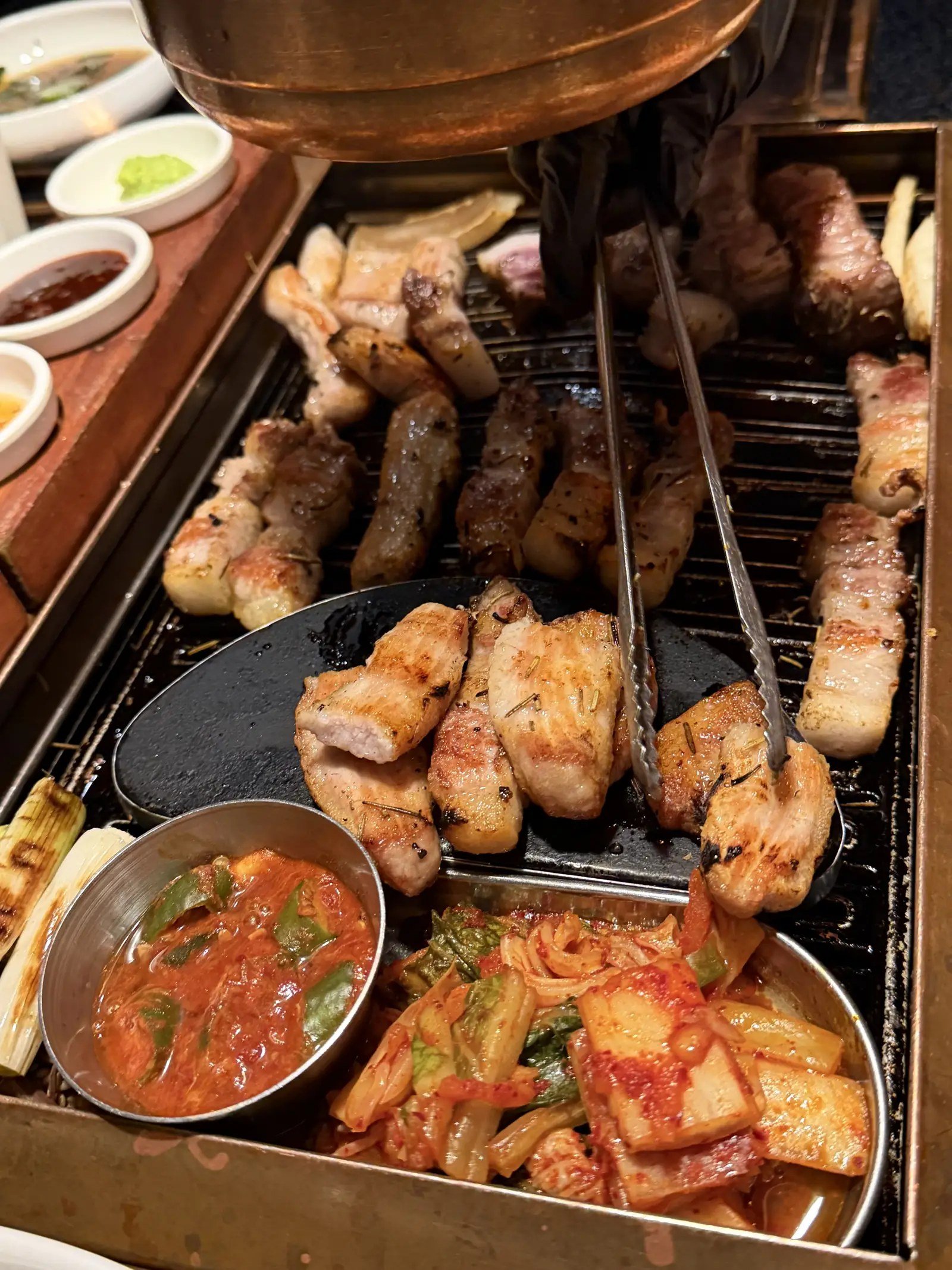 Tongs turning grilled pork on a Korean BBQ grill with a ventilation hood overhead.
