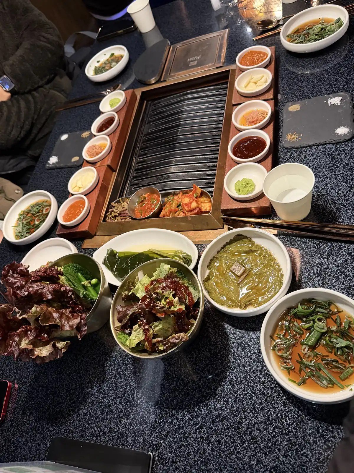 A Korean BBQ table with a built-in grill surrounded by banchan, lettuce, and dipping sauces.