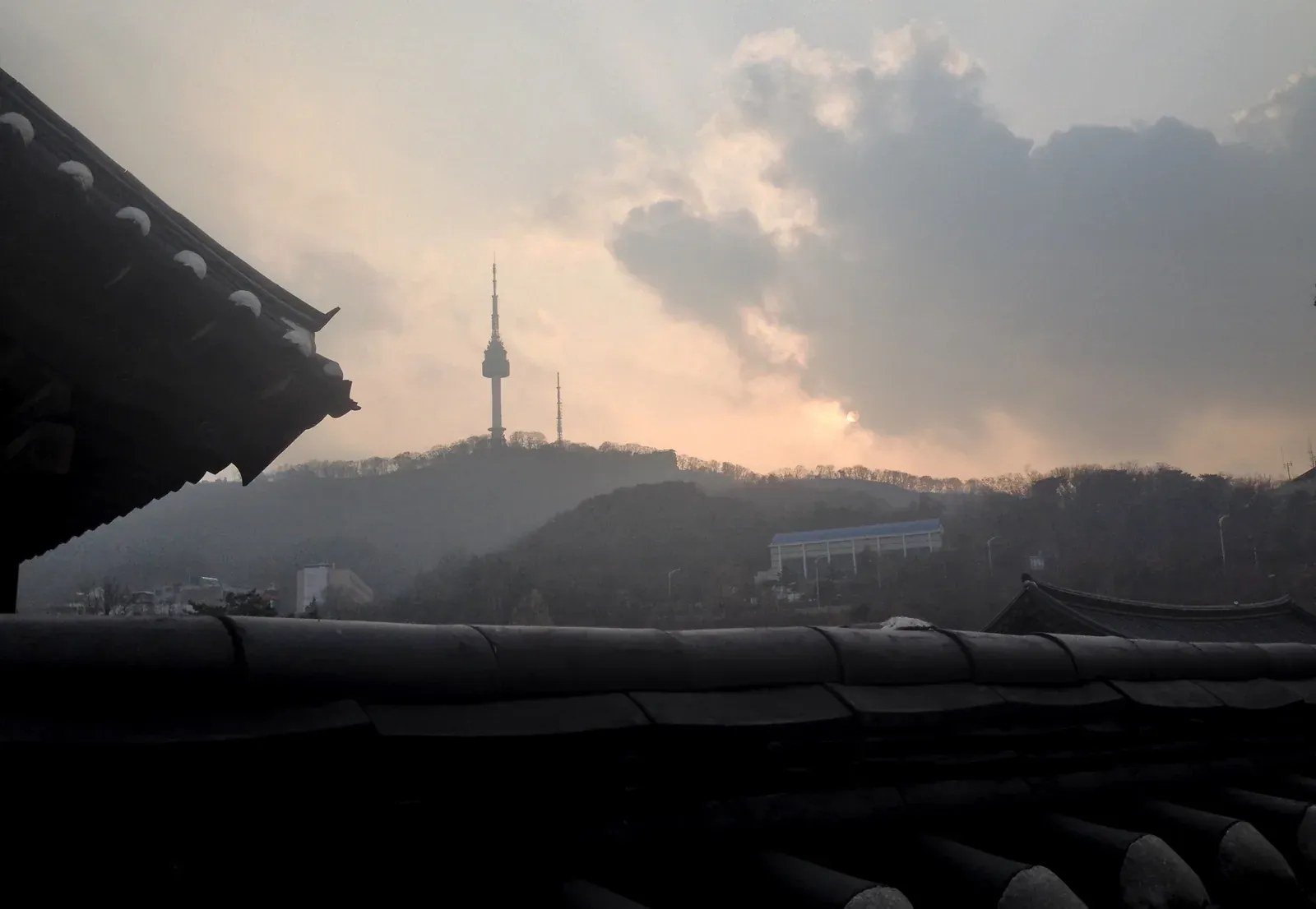 Dark hanok rooftops at Namsangol Hanok Village frame N Seoul Tower beneath a cloudy late-afternoon sky.