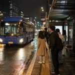 A blue city bus approaches a covered bus stop on a wet Seoul street at night while several adult passengers wait under streetlights.