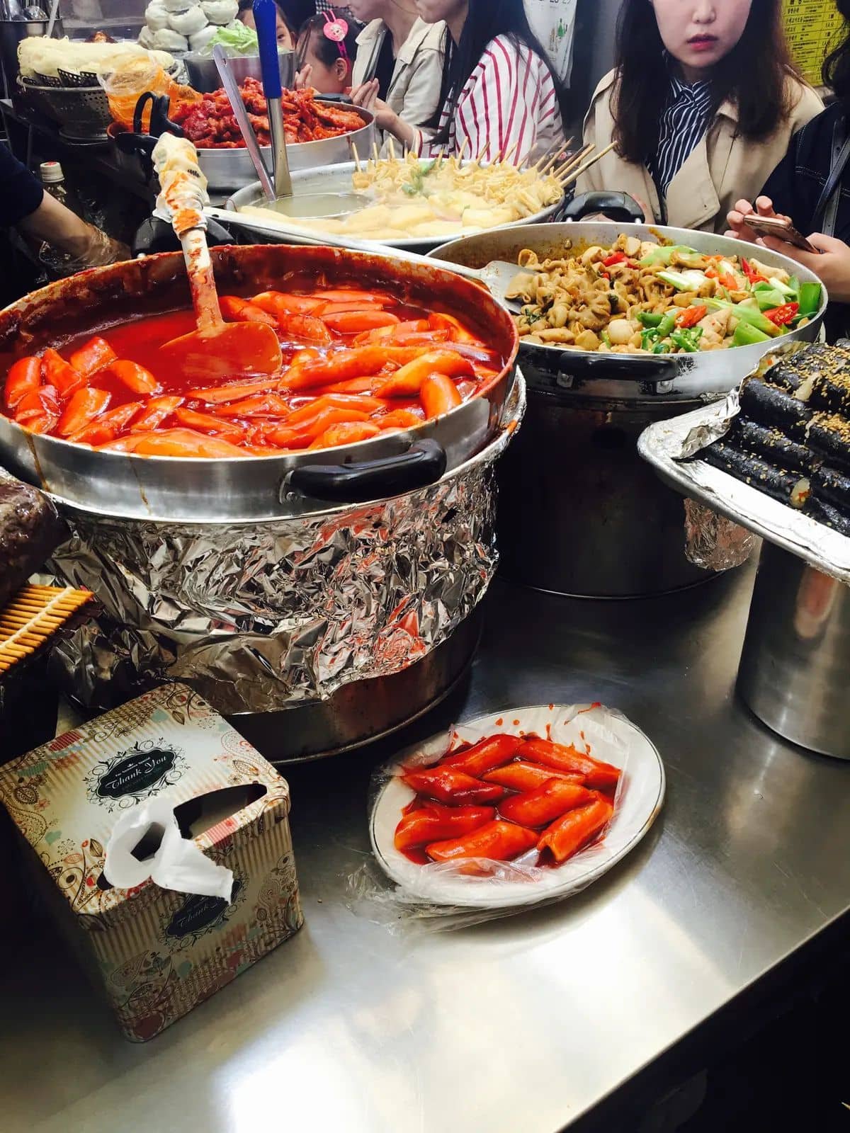 Busy Korean street-food counter with tteokbokki, fish cakes and diners.